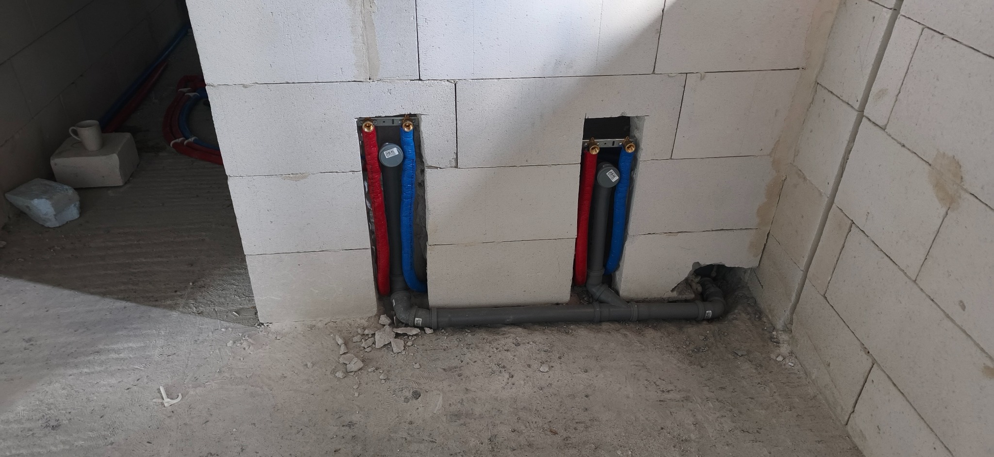 Instalacja wodna w ścianie z betonu komórkowego: czerwona i niebieska rura doprowadzona do podłączeń. Szare rury odpływowe. Widok z dołu na surowy stan.