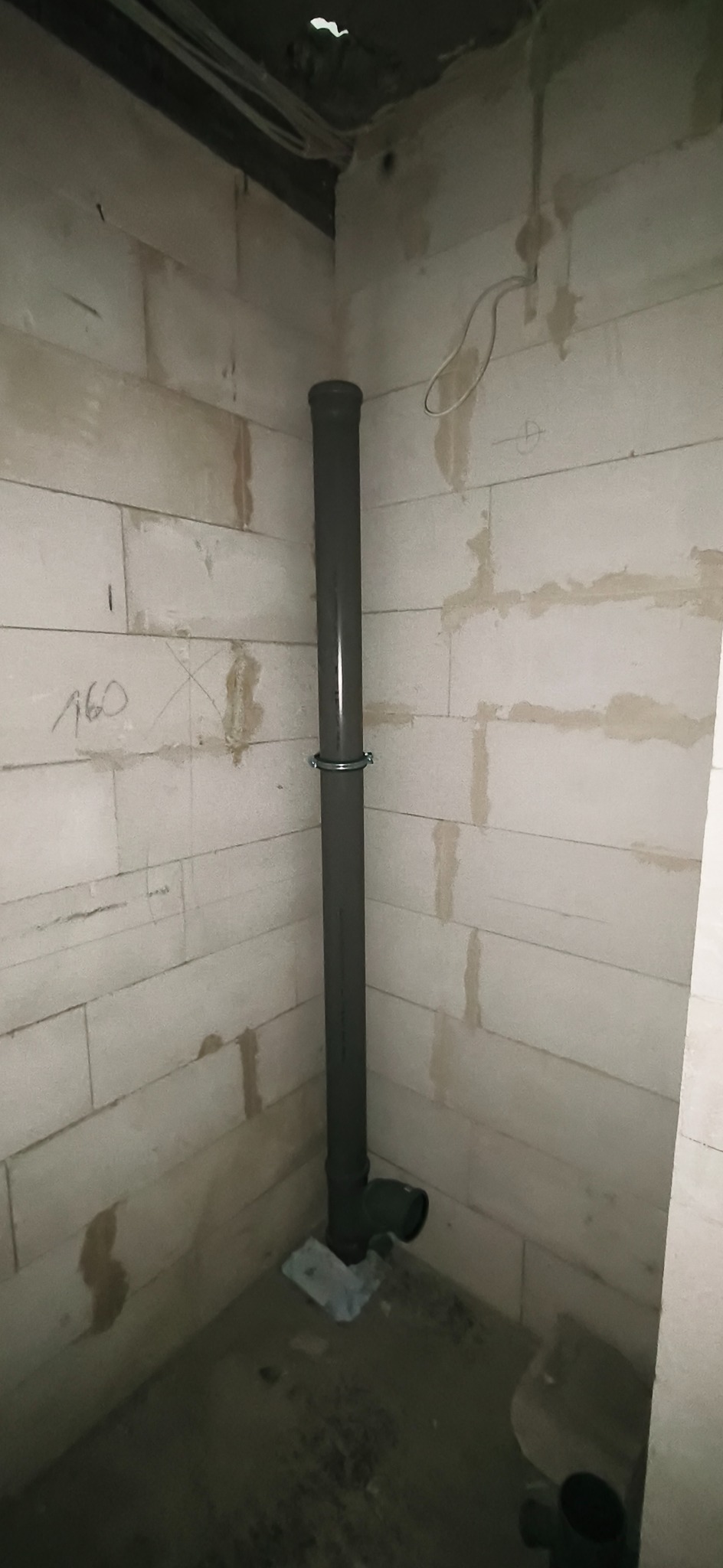 Szara rura kanalizacyjna pionowo zamocowana do ściany z betonu komórkowego za pomocą metalowej obejmy. Widoczne łączenie rur przy podłodze. Surowy stan wykończenia.