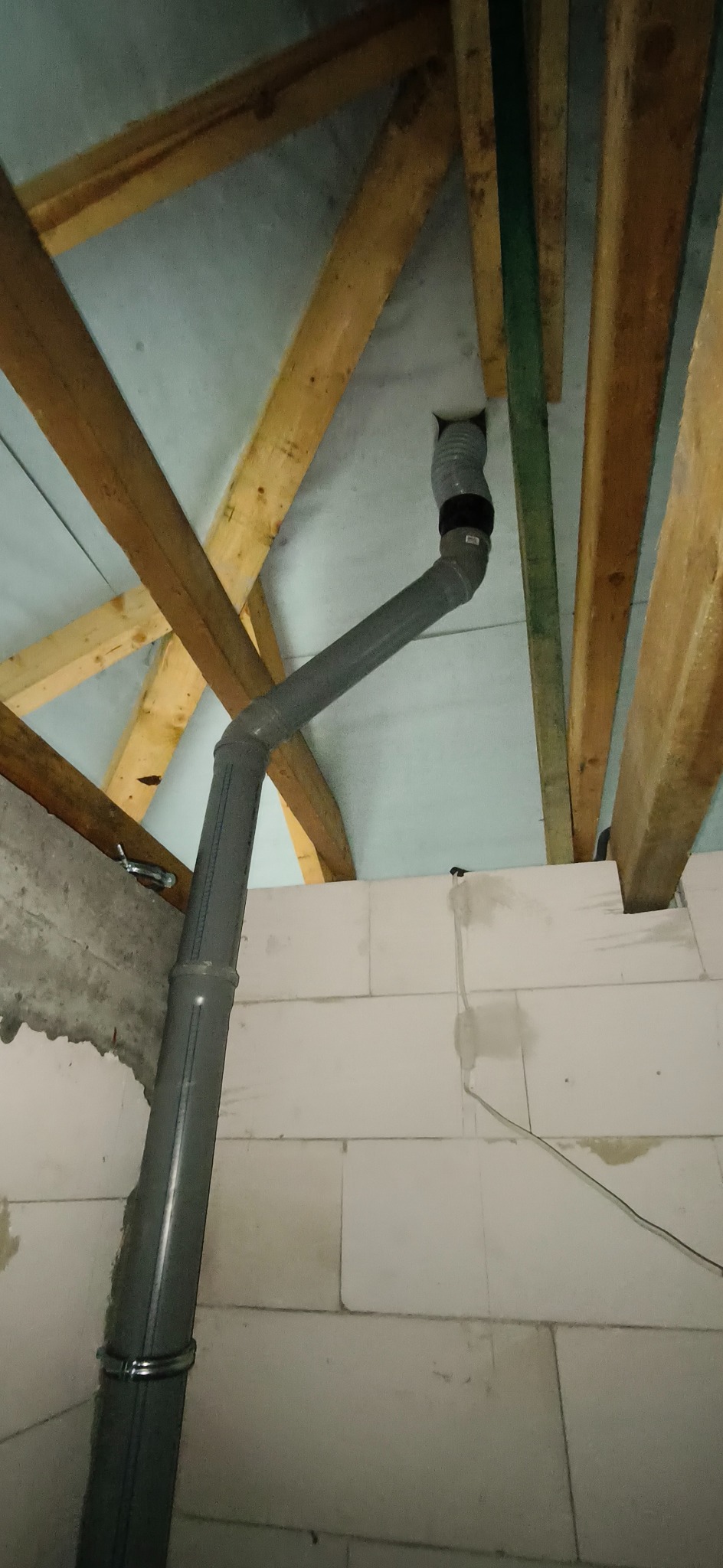 Szara rura kanalizacyjna z obejmą, biegnąca wzdłuż ściany z białych bloczków, w tle drewniana konstrukcja dachu. Widok z dołu, ukośny kadr.