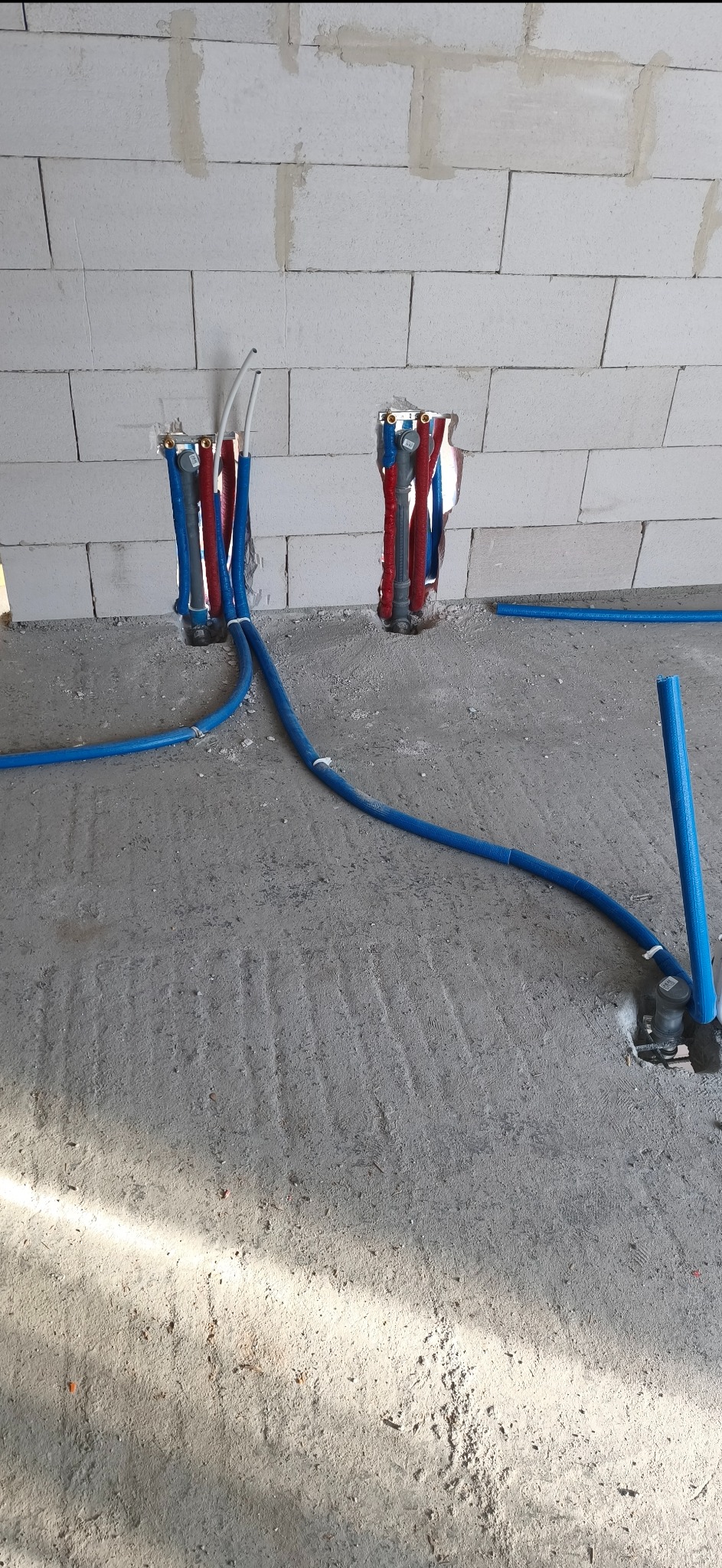 Instalacja wodna w ścianie z betonu komórkowego. Widoczne rury w kolorach niebieskim i czerwonym, prowadzone w podłodze. Etap prac instalacyjnych.