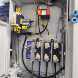 Rozdzielnia elektryczna w szarej obudowie z widocznymi elementami: bezpieczniki, przewody z oznaczeniami, złączki. Detal instalacji elektrycznej wewnątrz szafki.