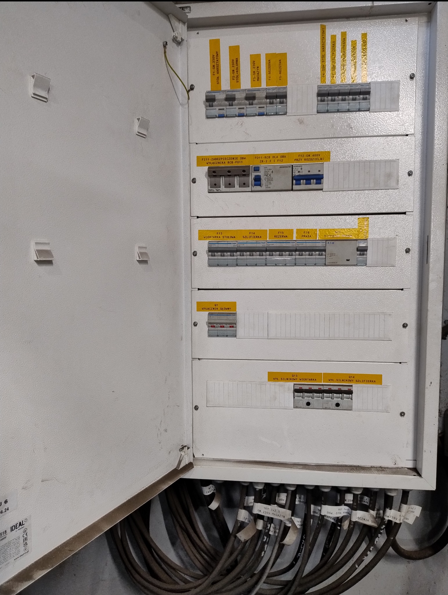 Otwarta skrzynka elektryczna z bezpiecznikami i opisami obwodów, widoczne kable zasilające, etykiety z oznaczeniami. Instalacja w kolorze białym, szare kable.