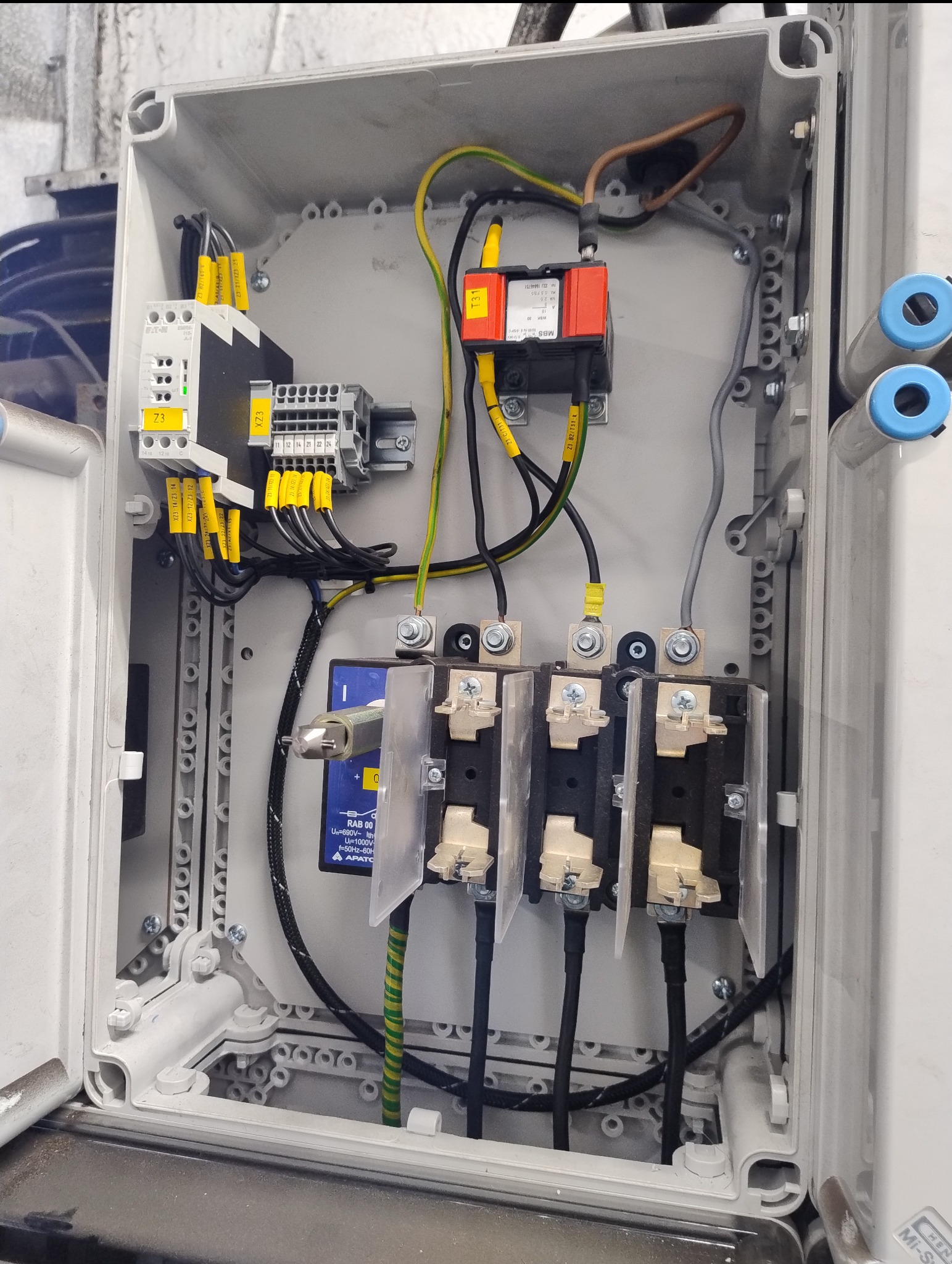 Rozdzielnia elektryczna w szarej obudowie z widocznymi elementami: bezpieczniki, przewody z oznaczeniami, złączki. Detal instalacji elektrycznej wewnątrz szafki.