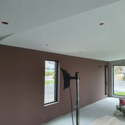 Oskar Ancypa - Świeżo pomalowane, brązowe ściany w pustym pokoju z dużymi oknami. Widoczne narzędzia malarskie, lampa oświetlająca ścianę. W tle widok na zewnątrz.