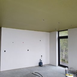 Oskar Ancypa - Wnętrze w trakcie wykańczania: białe ściany z otworami na gniazdka, oliwkowy sufit i duże okno z widokiem na zieleń. Widoczne narzędzia malarskie i sprzęt budowlany.