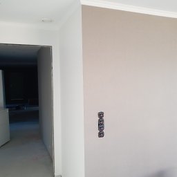 Oskar Ancypa - Jasna ściana z gładzią, ozdobnym gzymsem i trzema gniazdkami elektrycznymi w pionie. Widoczny fragment sufitu i wejście do pomieszczenia w głębi.