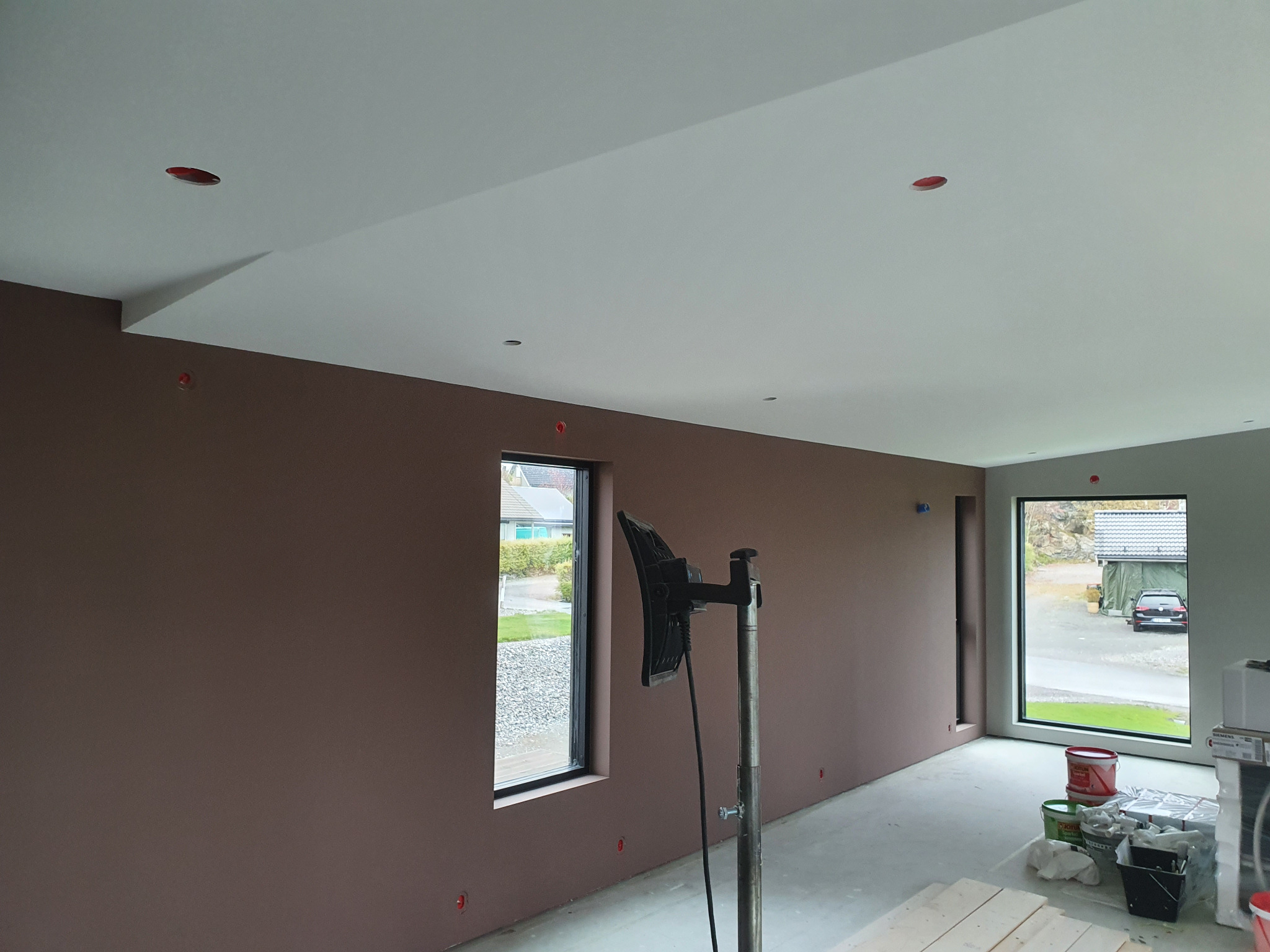 Świeżo pomalowane, brązowe ściany w pustym pokoju z dużymi oknami. Widoczne narzędzia malarskie, lampa oświetlająca ścianę. W tle widok na zewnątrz.