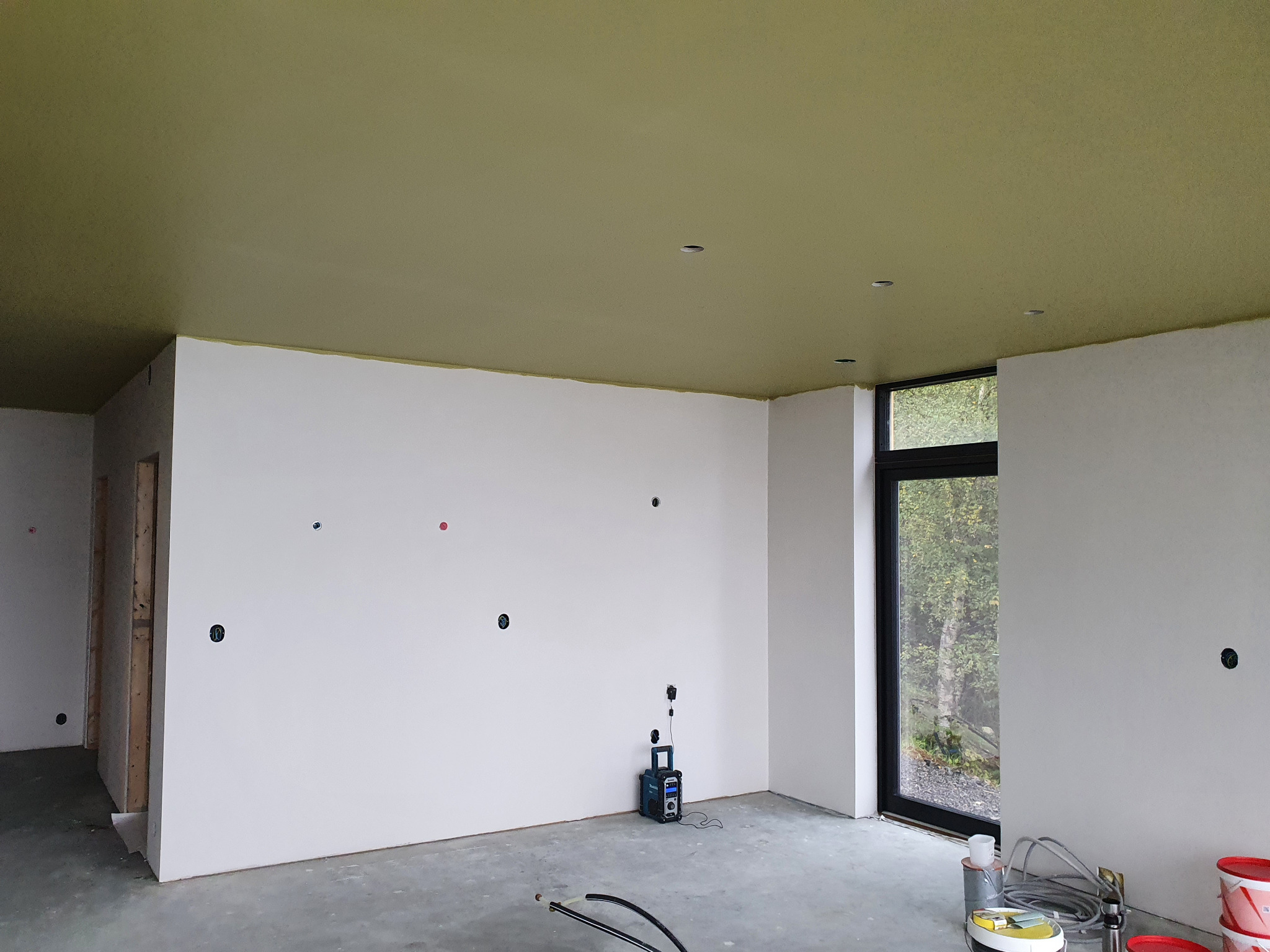 Wnętrze w trakcie wykańczania: białe ściany z otworami na gniazdka, oliwkowy sufit i duże okno z widokiem na zieleń. Widoczne narzędzia malarskie i sprzęt budowlany.