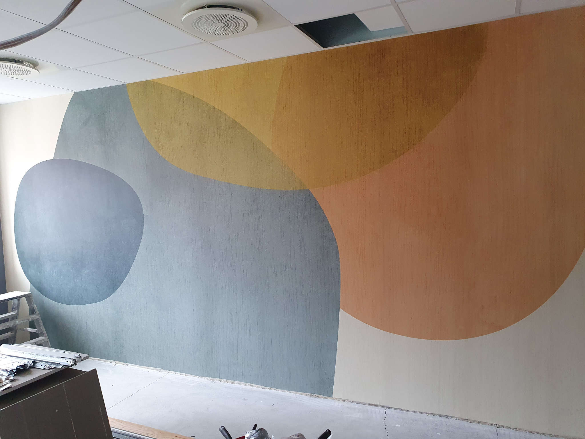 Ściana z abstrakcyjnym malowaniem w ciepłych i chłodnych odcieniach, widoczne drabina malarska i narzędzia, sugerujące prace wykończeniowe w pomieszczeniu.