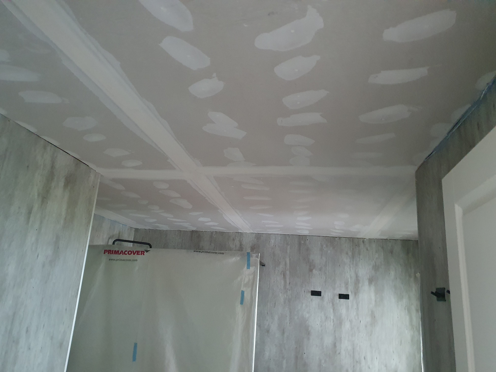 Sufit z płyt kartonowo-gipsowych, z widocznymi łączeniami i szpachlowaniem, przygotowany do dalszych prac wykończeniowych. Ściany surowe, zabezpieczone folią.