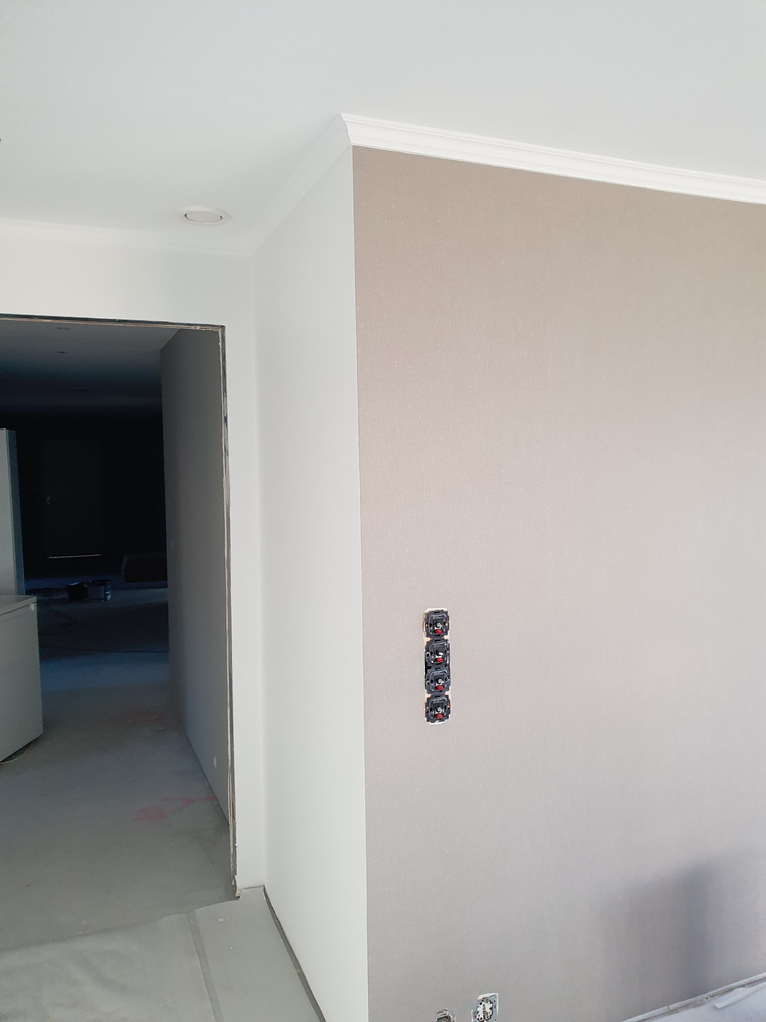 Jasna ściana z gładzią, ozdobnym gzymsem i trzema gniazdkami elektrycznymi w pionie. Widoczny fragment sufitu i wejście do pomieszczenia w głębi.