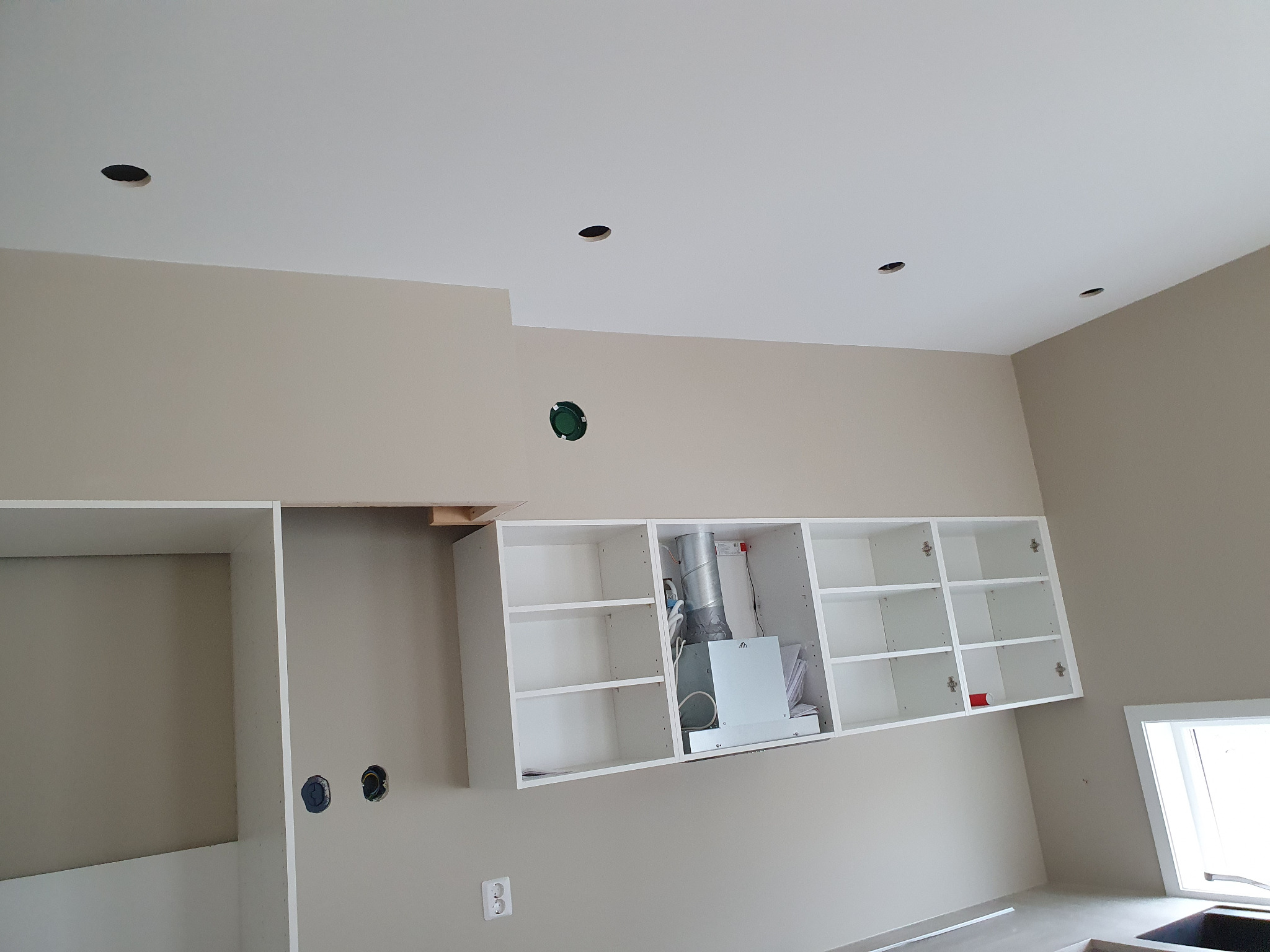 Wnętrze kuchni w trakcie montażu: białe szafki wiszące bez frontów, otwory w suficie pod oświetlenie, jasne ściany. Widoczne elementy instalacji i okno.