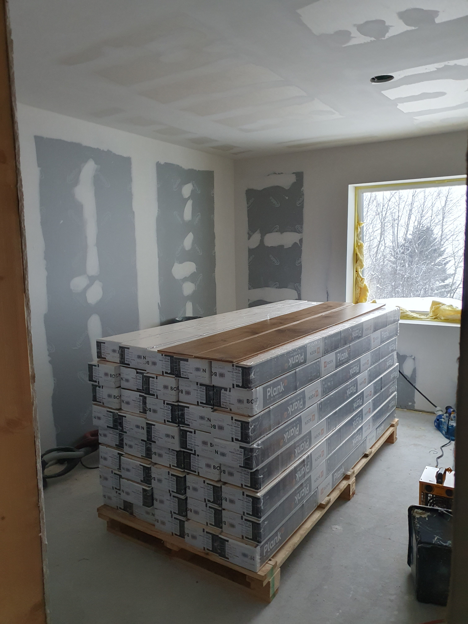 Stos paneli podłogowych na palecie w surowym wnętrzu. Ściany ze śladami szpachlowania, okno z widokiem na zimowy krajobraz. Przygotowanie do układania paneli.
