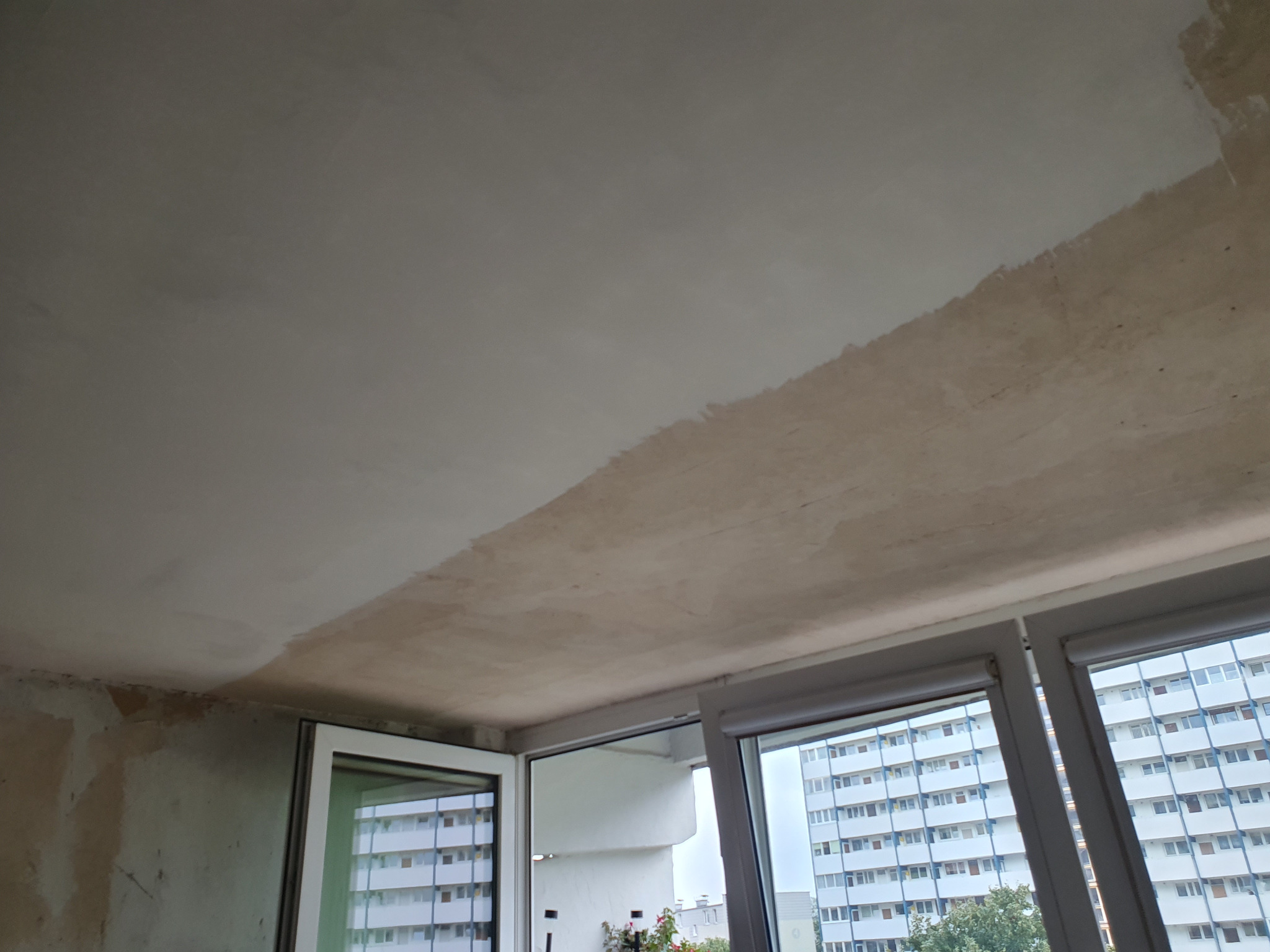 Częściowo wyszpachlowany sufit w mieszkaniu, widoczne okna z widokiem na blok. Fragment ściany z pozostałościami po starych powłokach. Etap prac wykończeniowych.