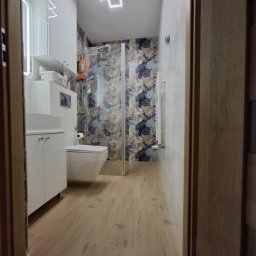 Remonty Na Poziomie Filip Kitliński Usługi Wykończeniowo-Remontowe - Nowoczesna łazienka po remoncie: kabina prysznicowa ze szklaną ścianą, umywalka z szafką, toaleta w zabudowie, grzejnik drabinkowy i podświetlane lustro.