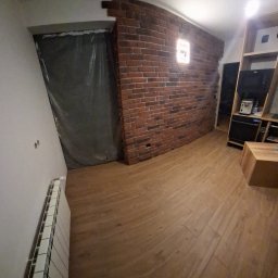 Remonty Na Poziomie Filip Kitliński Usługi Wykończeniowo-Remontowe - Kuchnia po remoncie: ceglana ściana z oświetleniem, drewnopodobne płytki na podłodze, zabudowa kuchenna. Część pomieszczenia zabezpieczona folią.