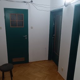 Remonty Na Poziomie Filip Kitliński Usługi Wykończeniowo-Remontowe - Korytarz po remoncie: świeżo pomalowane ściany na biało, odnowiony parkiet w jodełkę, ciemnozielone drzwi z szybką i krzesło. Widoczne kable pod sufitem.