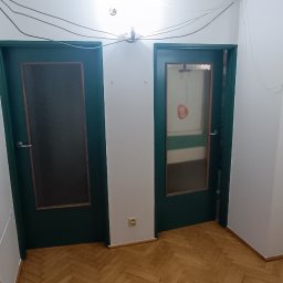 Remonty Na Poziomie Filip Kitliński Usługi Wykończeniowo-Remontowe - Korytarz po remoncie: odnowione ściany, pomalowane na biało, trzy zielone drzwi z szybami, parkiet na podłodze, widoczne kable pod sufitem.