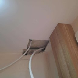 Remonty Na Poziomie Filip Kitliński Usługi Wykończeniowo-Remontowe - Otwór w suficie podwieszanym z widocznymi rurami, przygotowany do naprawy. Widoczne uszkodzenia i fragment drewnianej konstrukcji. Ujęcie z dołu.