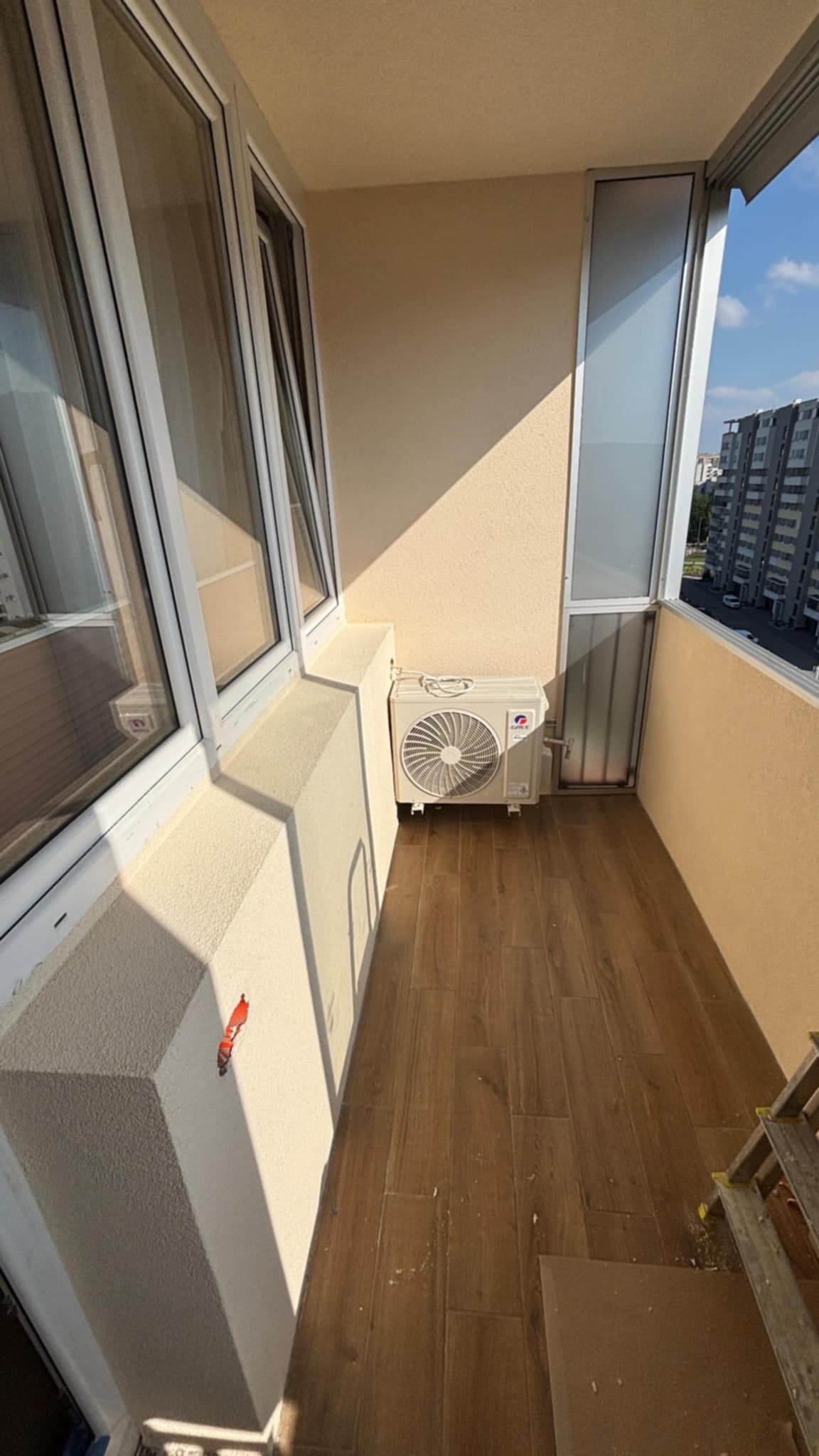 Wykończony balkon z drewnopodobnymi płytkami podłogowymi, klimatyzatorem i świeżo otynkowanymi ścianami w beżowym odcieniu, widok z góry na zewnątrz.