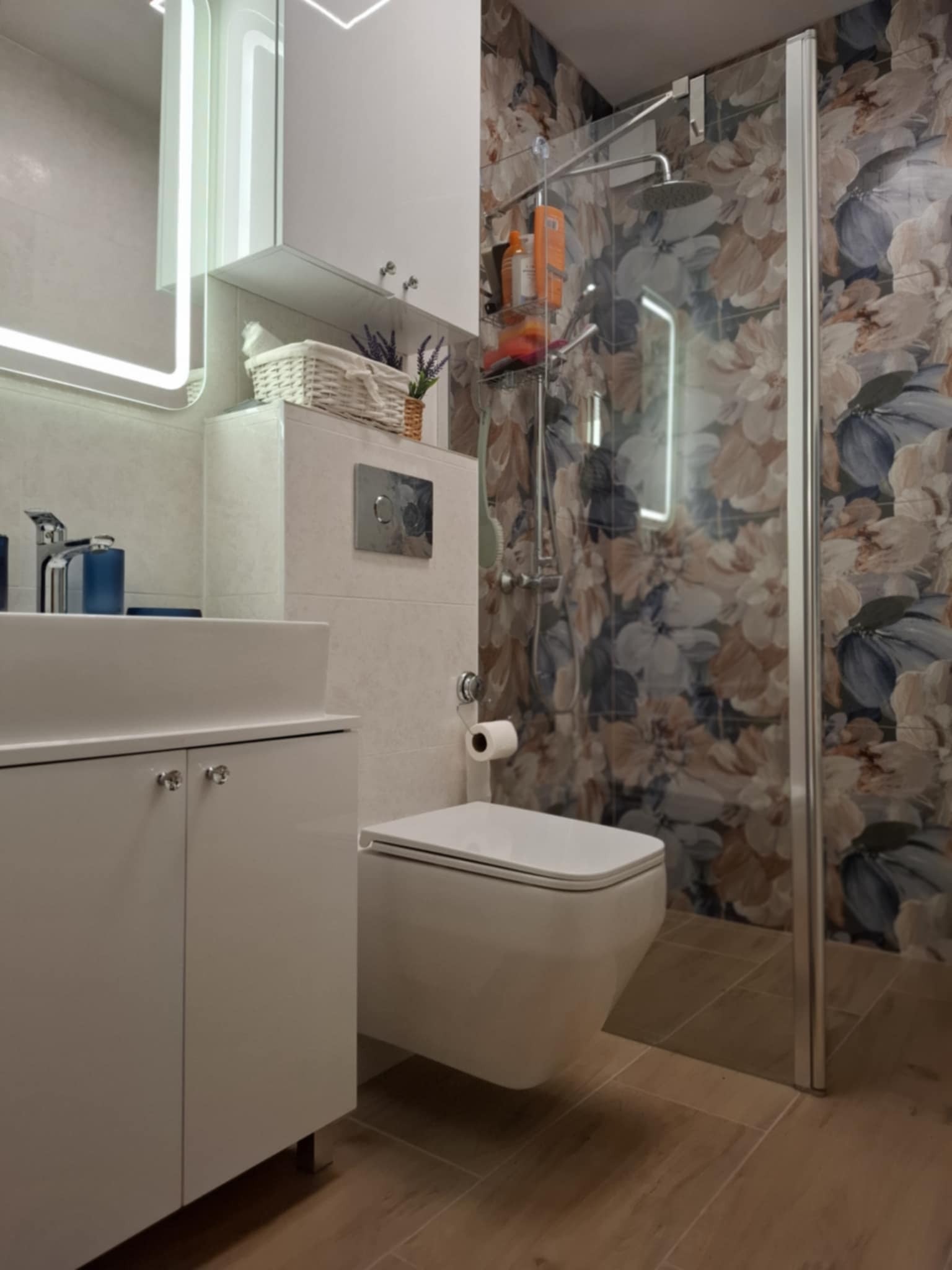 Nowoczesna łazienka z kabiną prysznicową w kwiaty, umywalką na szafce i podświetlanym lustrem. Uwagę zwracają drewnopodobne płytki podłogowe i armatura.