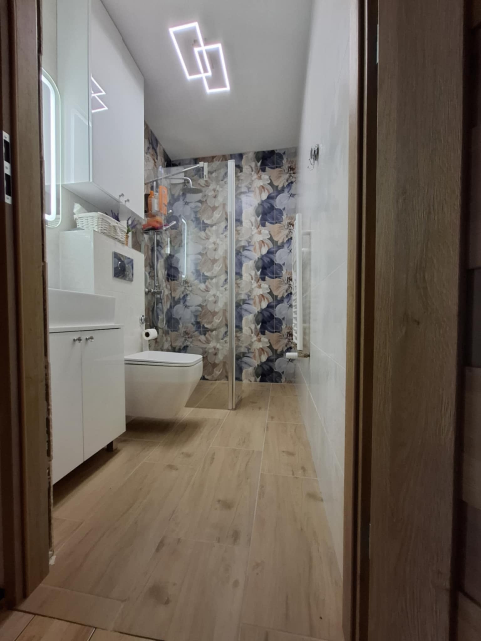 Nowoczesna łazienka po remoncie: kabina prysznicowa ze szklaną ścianą, umywalka z szafką, toaleta w zabudowie, grzejnik drabinkowy i podświetlane lustro.