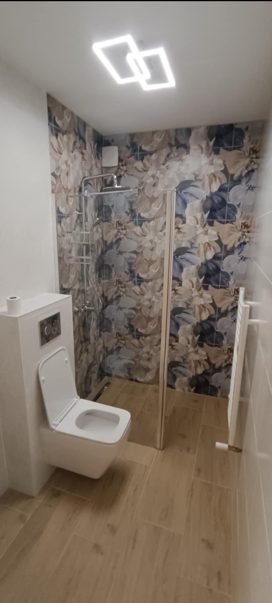 Nowoczesna łazienka po remoncie: kabina prysznicowa ze szklaną ścianką, podwieszana toaleta, grzejnik drabinkowy i płytki z motywem kwiatowym.