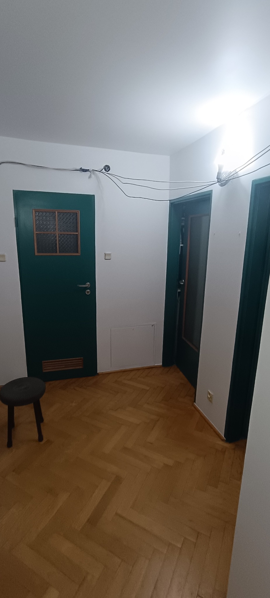Korytarz po remoncie: świeżo pomalowane ściany na biało, odnowiony parkiet w jodełkę, ciemnozielone drzwi z szybką i krzesło. Widoczne kable pod sufitem.