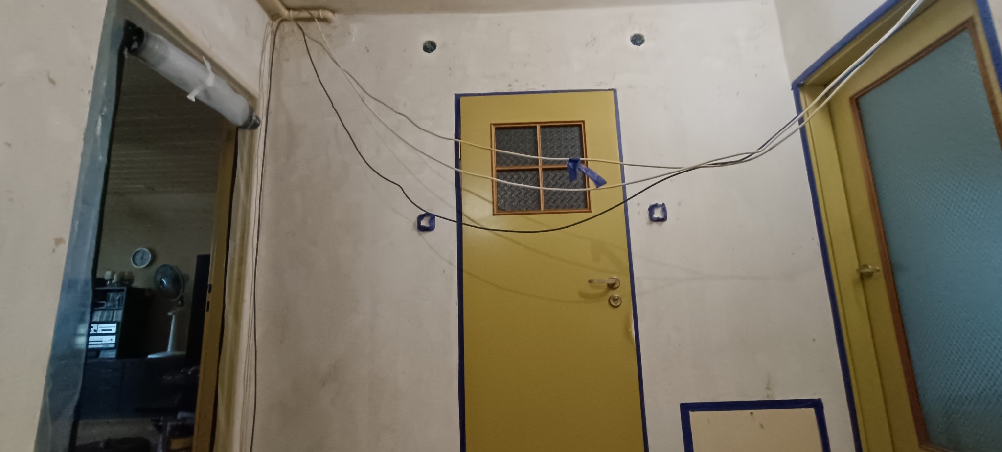 Korytarz w trakcie remontu: ściany z ubytkami, widoczne kable, żółte drzwi z niebieską taśmą. Ujęcie z dołu, widok na wnętrze przez otwarte drzwi.