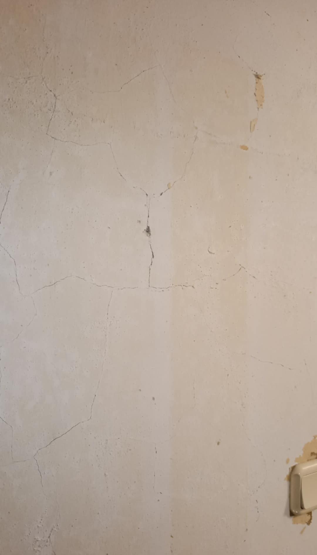 Ściana w odcieniach beżu z widocznymi pęknięciami i ubytkami tynku, fragment włącznika światła w prawym dolnym rogu. Stan 'przed' remontem.