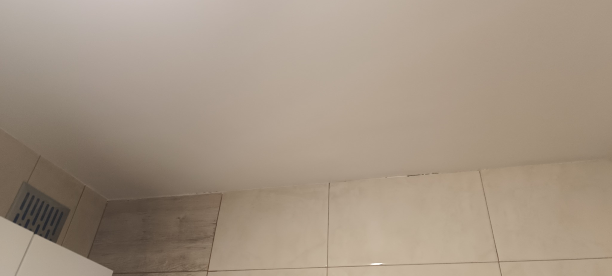 Wykończony sufit podwieszany po naprawie dziur, z widoczną kratką wentylacyjną i fragmentem ściany wyłożonej płytkami w odcieniach beżu i szarości. Gładka powierzchnia sufitu.