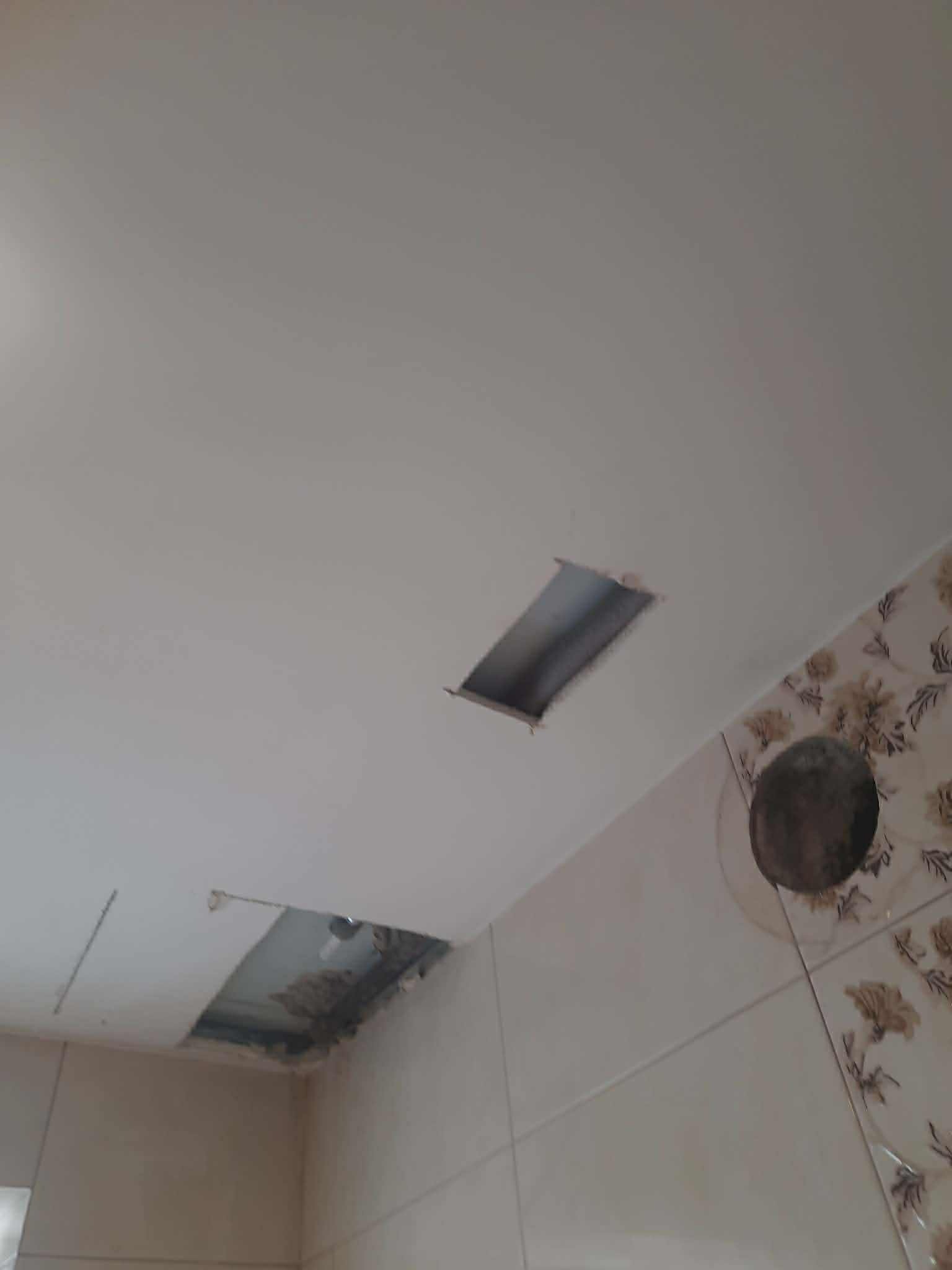 Otwory w suficie podwieszanym w łazience, widoczne rury. Przygotowanie do naprawy i szpachlowania. Fragment ściany z kwiatowym wzorem. Ujęcie z dołu.