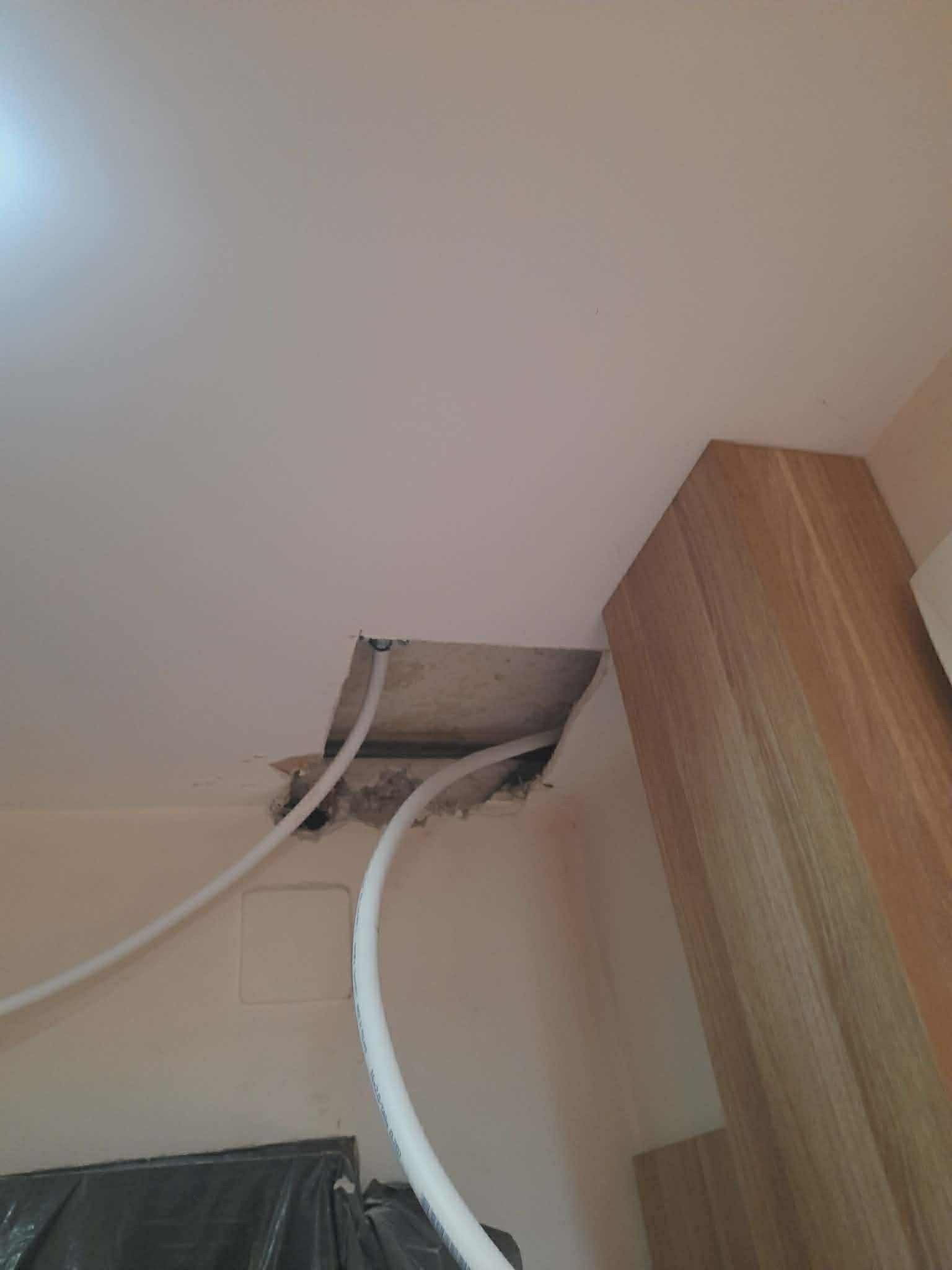 Otwór w suficie podwieszanym z widocznymi rurami, przygotowany do naprawy. Widoczne uszkodzenia i fragment drewnianej konstrukcji. Ujęcie z dołu.