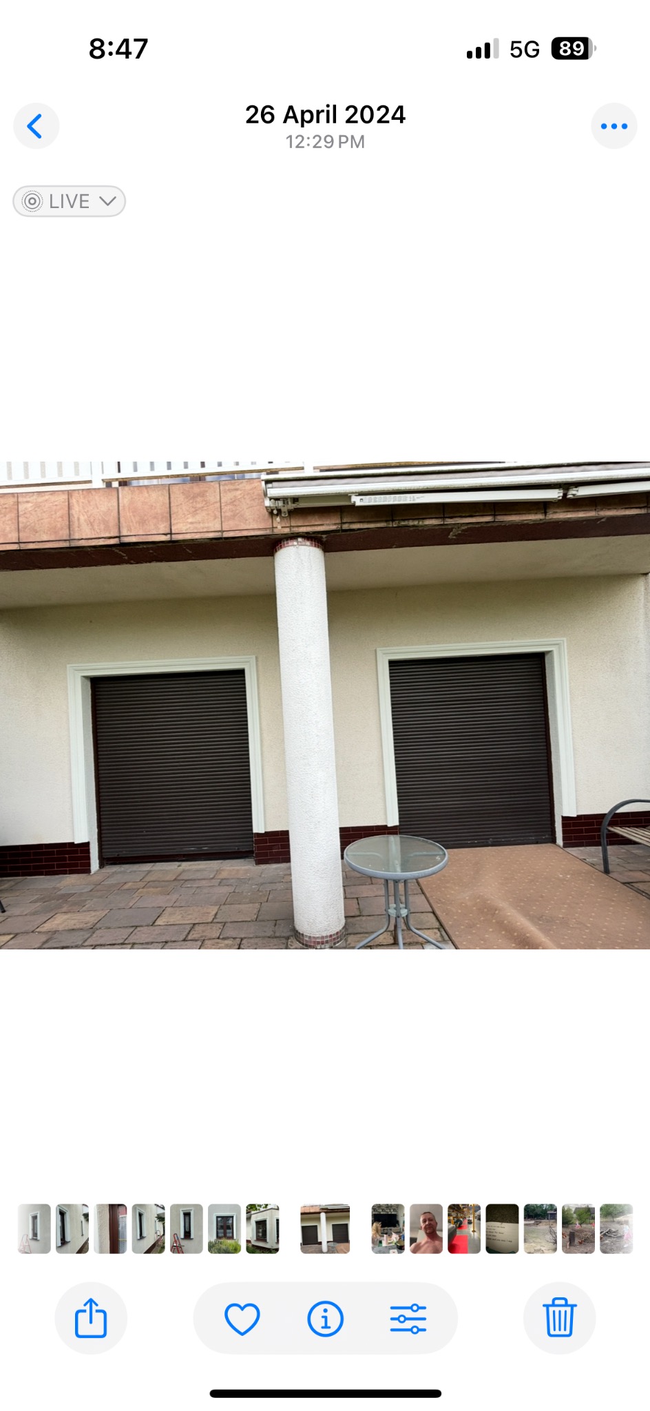 Zewnętrzny widok budynku z brązowymi roletami, kolumną i stolikiem. Widoczny fragment wyłożonego kostką podwórka. Ujęcie z poziomu gruntu, lekko z dołu.