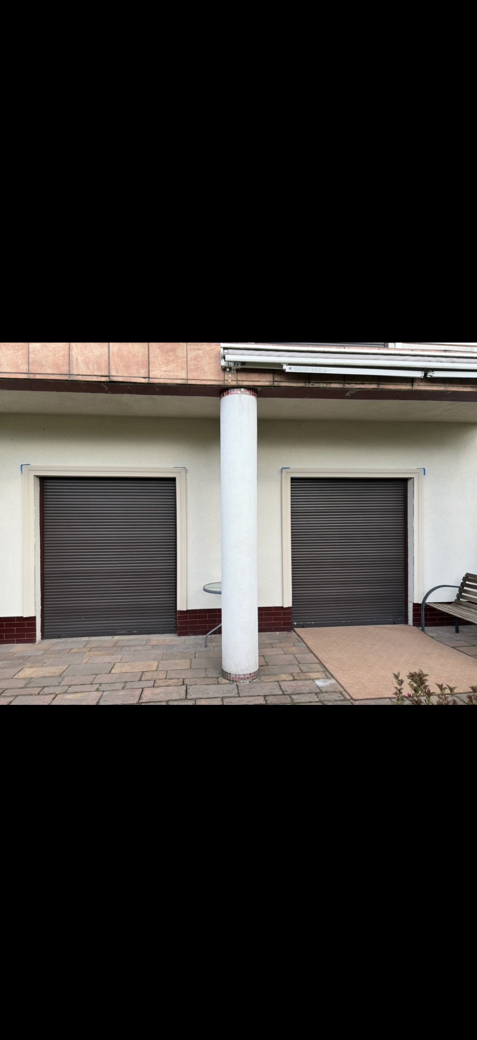 Elewacja budynku z dwoma oknami z brązowymi roletami, białą kolumną i fragmentem chodnika z kostki brukowej. Widoczna ławka i mały stolik.