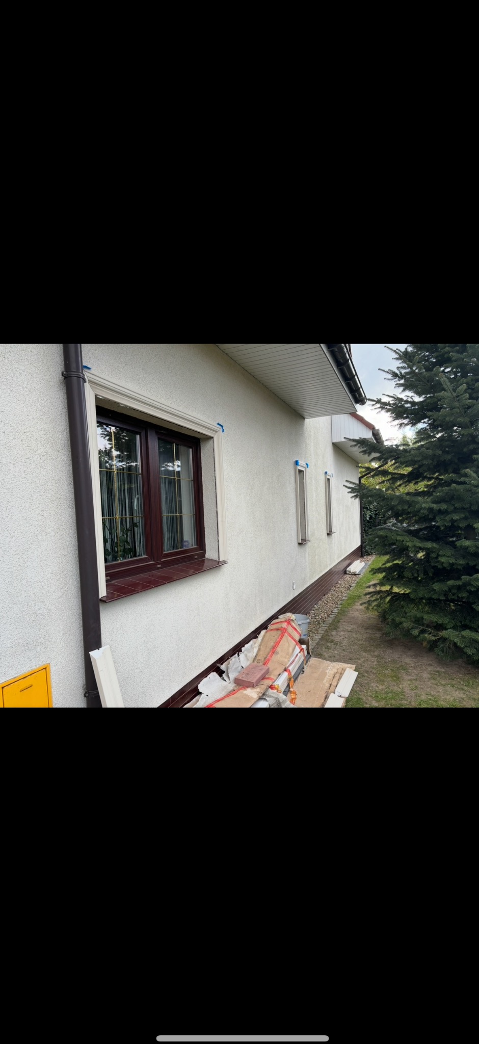 Elewacja domu jednorodzinnego z nową stolarką okienną w brązowych ramach, z zamontowanymi białymi listwami dekoracyjnymi. Na ziemi leżą materiały budowlane.