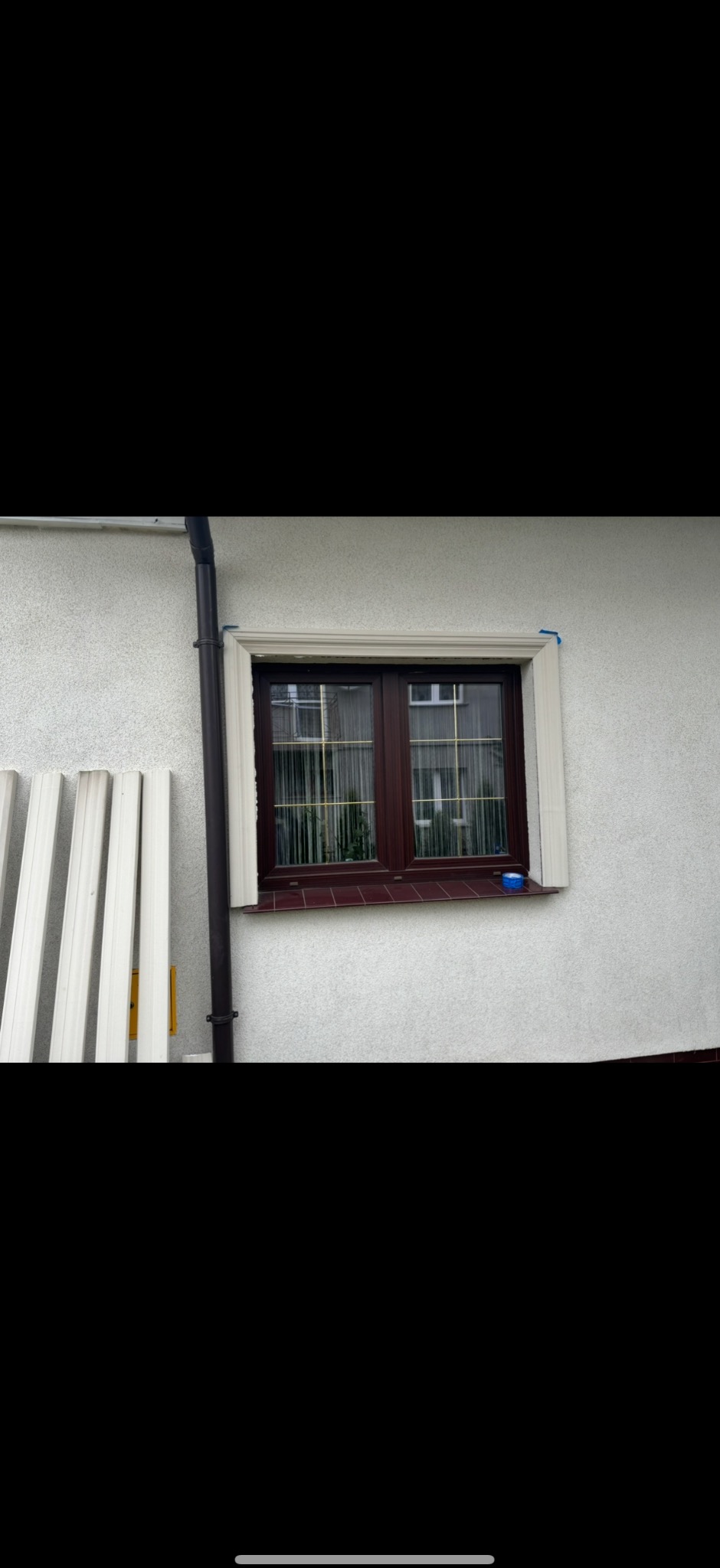 Okna z brązową ramą, białą listwą i parapetem z płytek. Na ścianie widoczny fragment rynny i przygotowane białe panele. Detal: niebieska taśma malarska na listwie.