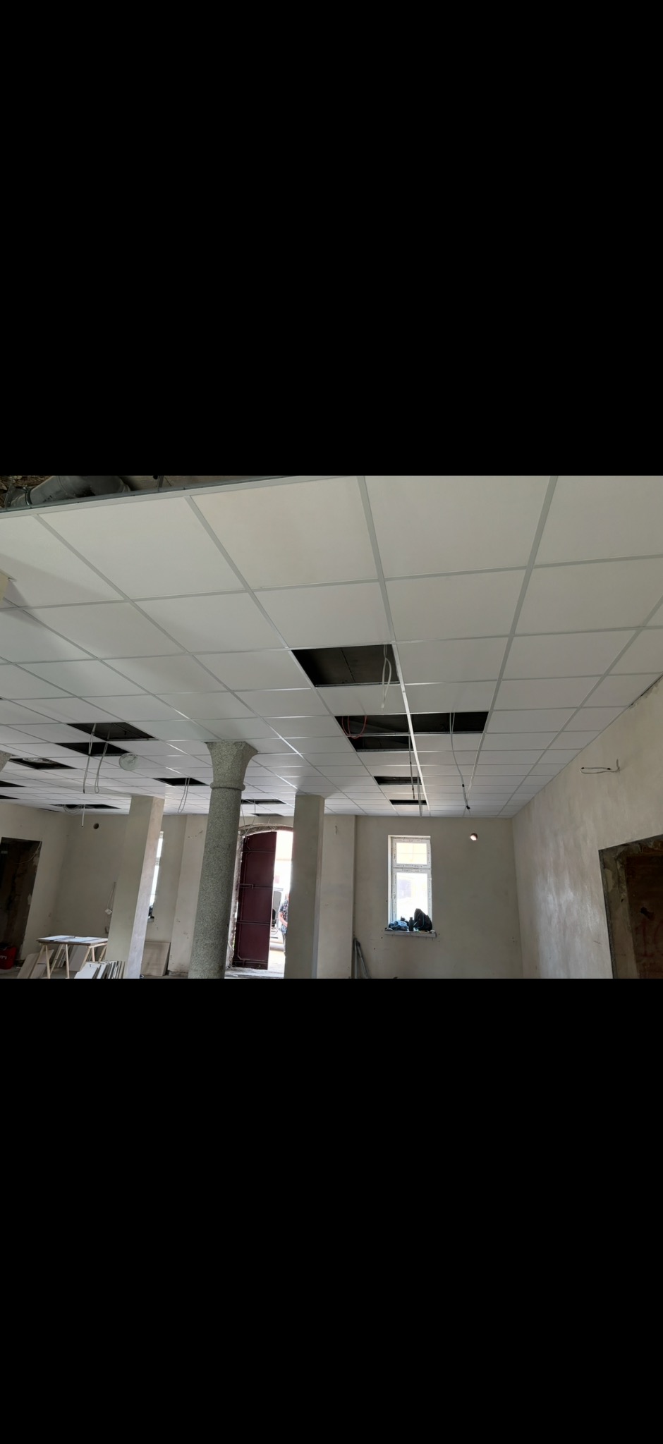Sufit podwieszany kasetonowy w trakcie montażu, widoczne kolumny i okno w tle. Część kasetonów brakuje, odsłaniając instalacje.