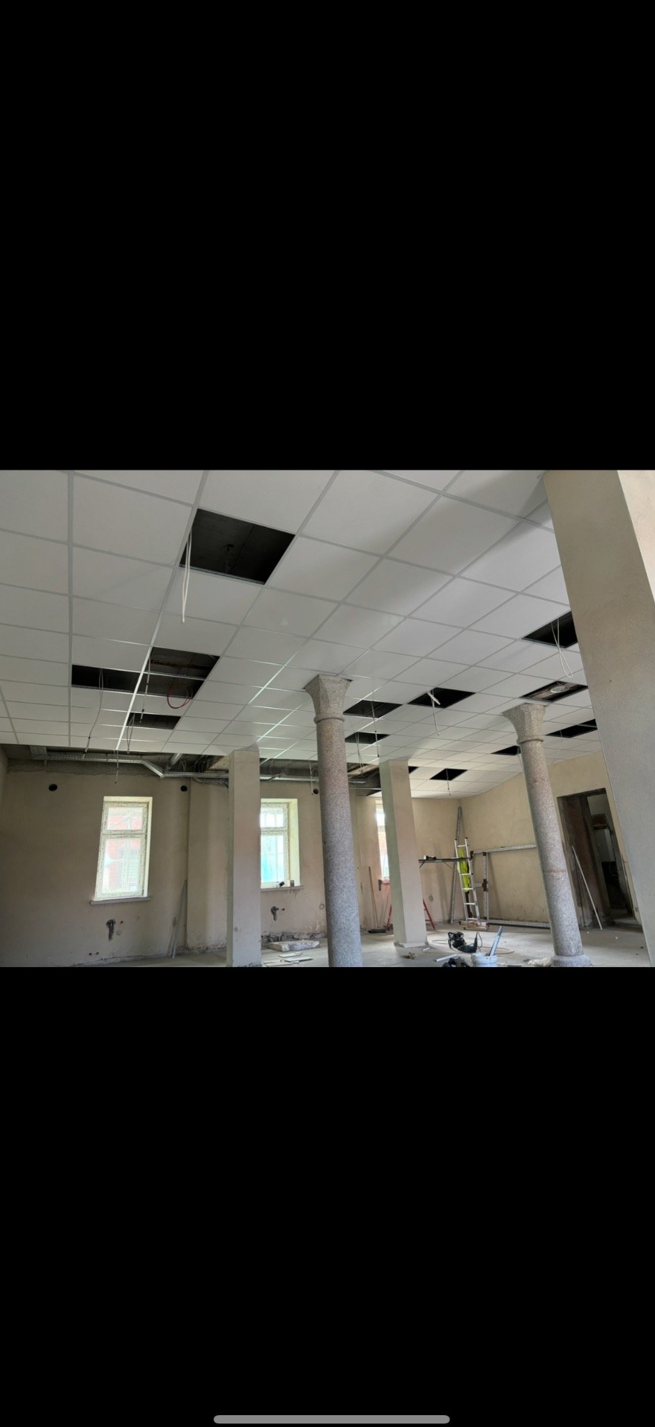 Wnętrze w trakcie prac wykończeniowych: sufit kasetonowy z otworami, kolumny z dekoracyjnym wykończeniem, ściany przygotowane do malowania, drabina w tle.