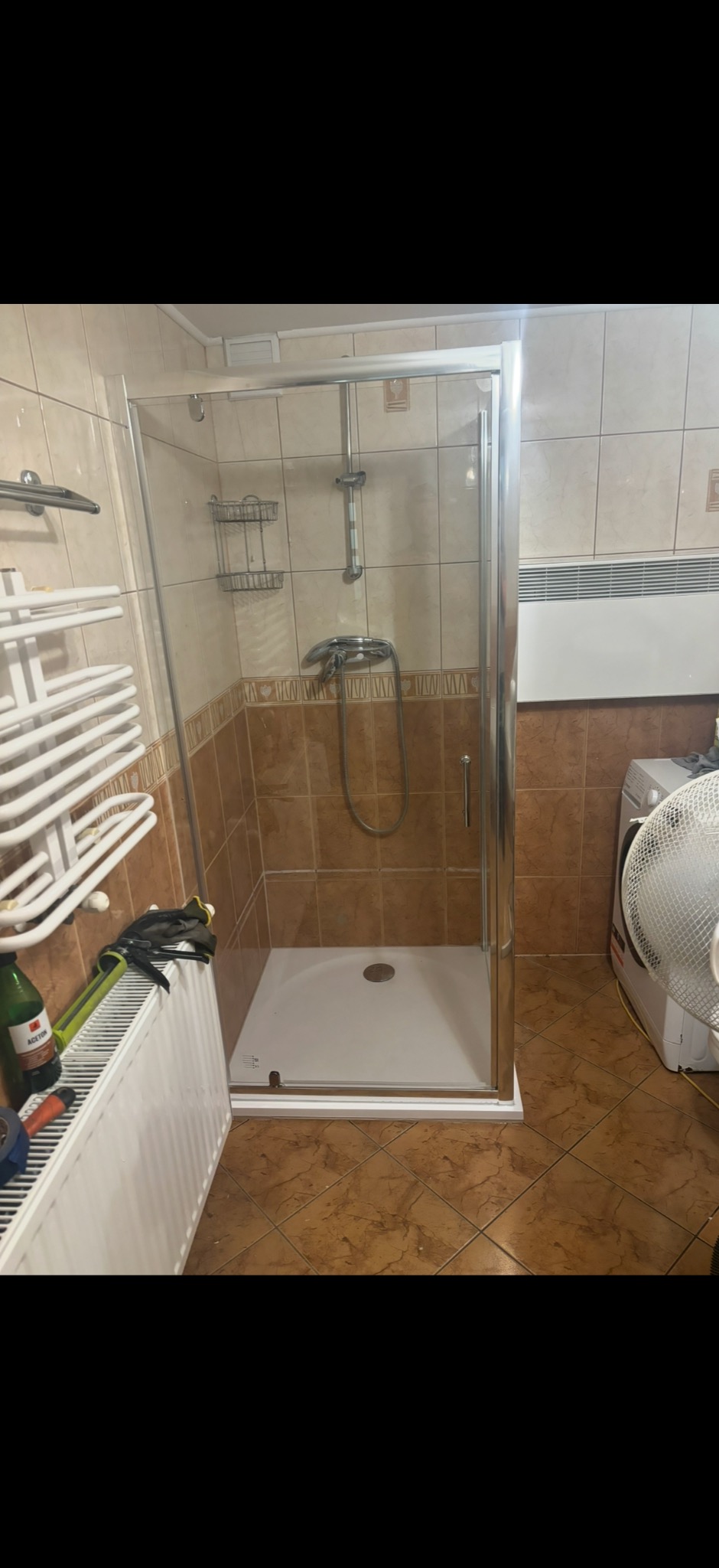 Zamontowana kabina prysznicowa z chromowanymi profilami i białym brodzikiem w łazience wykończonej brązowymi płytkami. Widoczny grzejnik i wentylator.