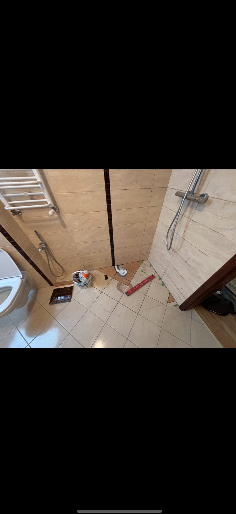 Remont łazienki w toku: częściowo ułożone beżowe płytki na podłodze, poziomica, wiadro z zaprawą. Fragment toalety i grzejnika w tle.