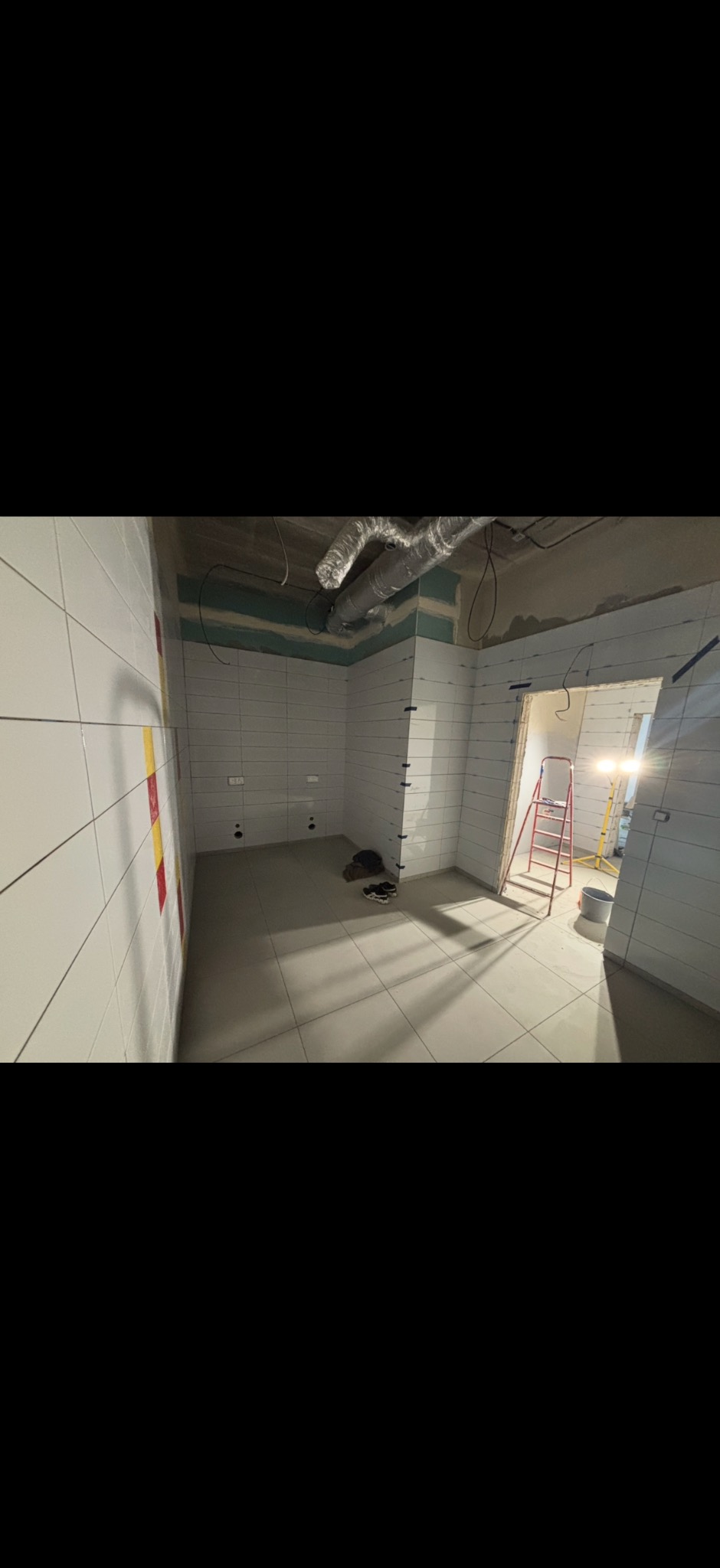 Wnętrze w trakcie prac wykończeniowych: białe płytki na ścianach i podłodze, widoczna drabina, instalacja wentylacyjna pod sufitem. W tle widać światło z zewnątrz.