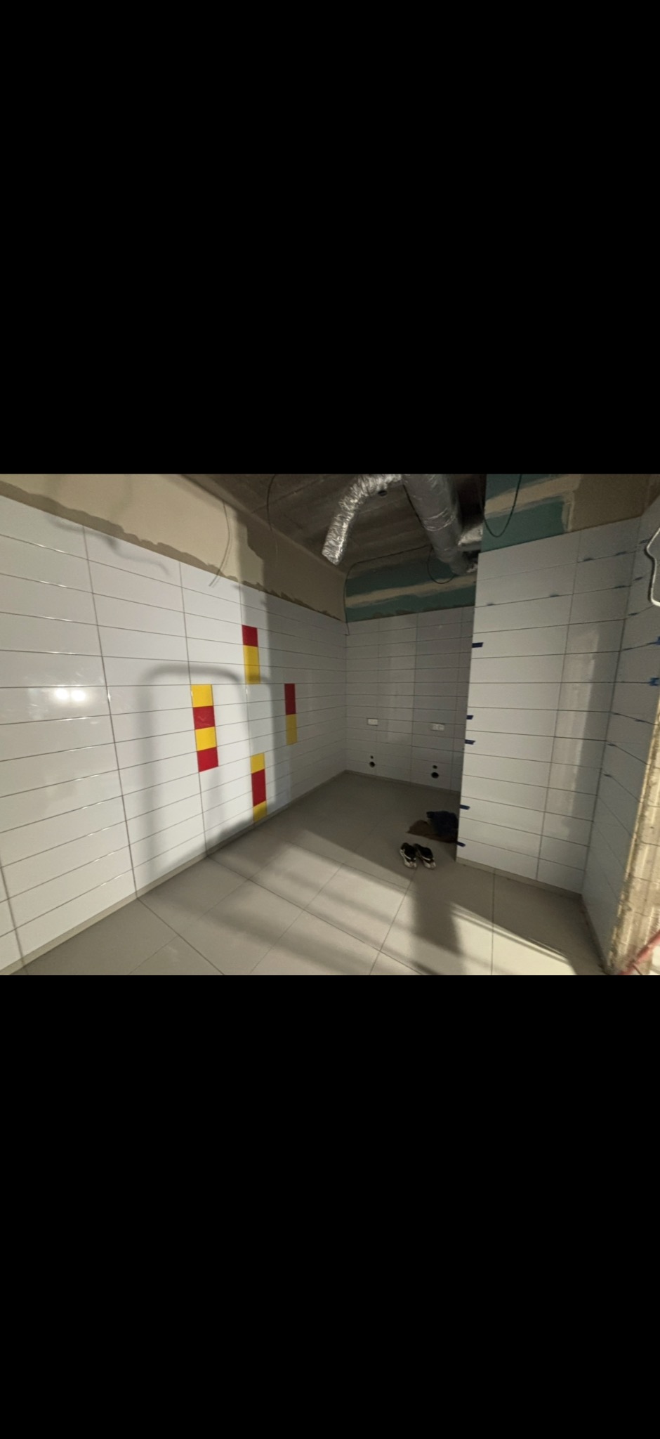 Wnętrze w trakcie prac wykończeniowych: białe płytki na ścianach z akcentami żółto-czerwonymi, jasna podłoga. Widoczne elementy instalacji pod sufitem.