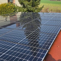 Pod Ciśnieniem - Marcin Piesiur - Błyszczące panele fotowoltaiczne na czerwonym dachu, odbijające budynek i drzewa, z narzędziem do czyszczenia paneli obok.