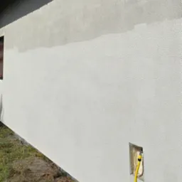 Ściana budynku z nową, jasną elewacją. Widoczny fragment okna, trawa przy budynku i żółty wąż w otworze w ścianie.