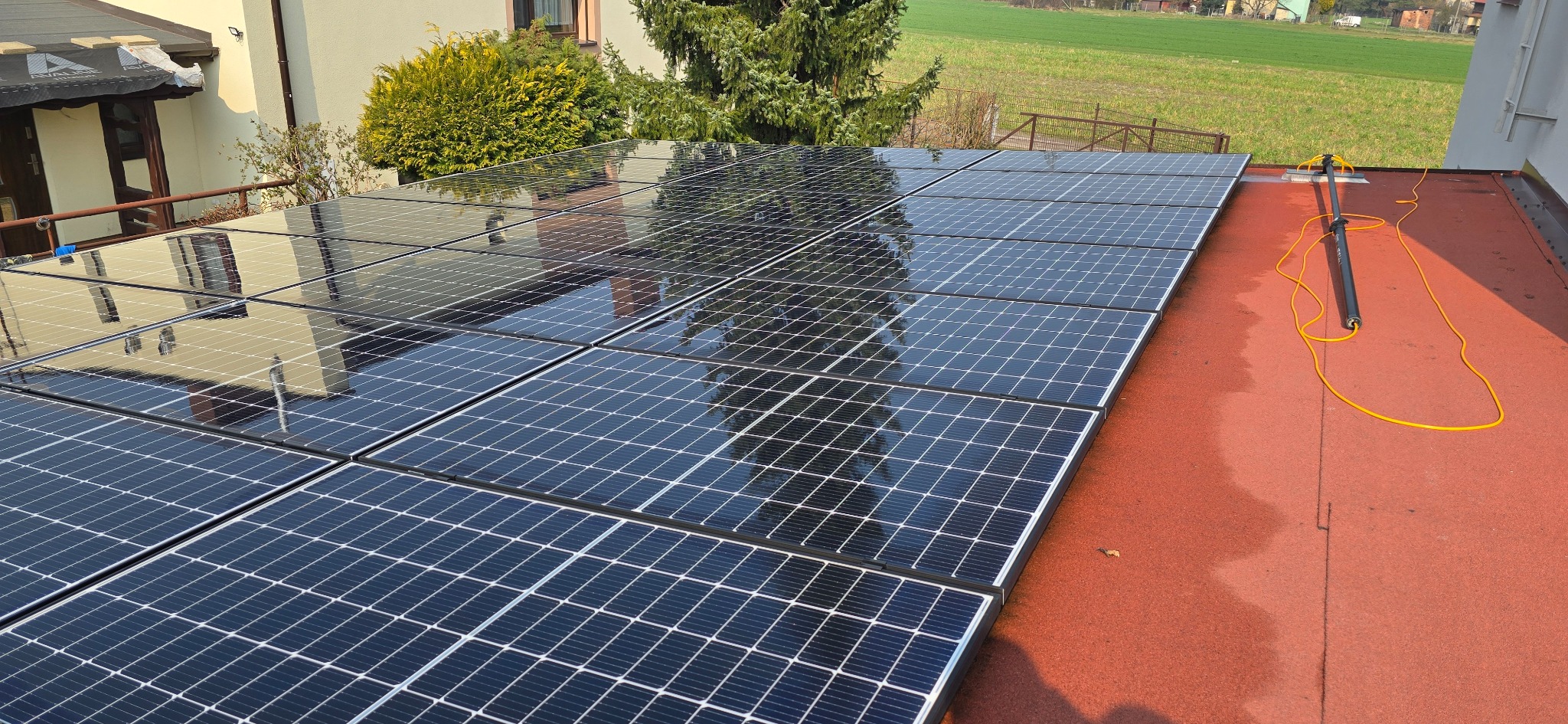 Błyszczące panele fotowoltaiczne na czerwonym dachu, odbijające budynek i drzewa, z narzędziem do czyszczenia paneli obok.