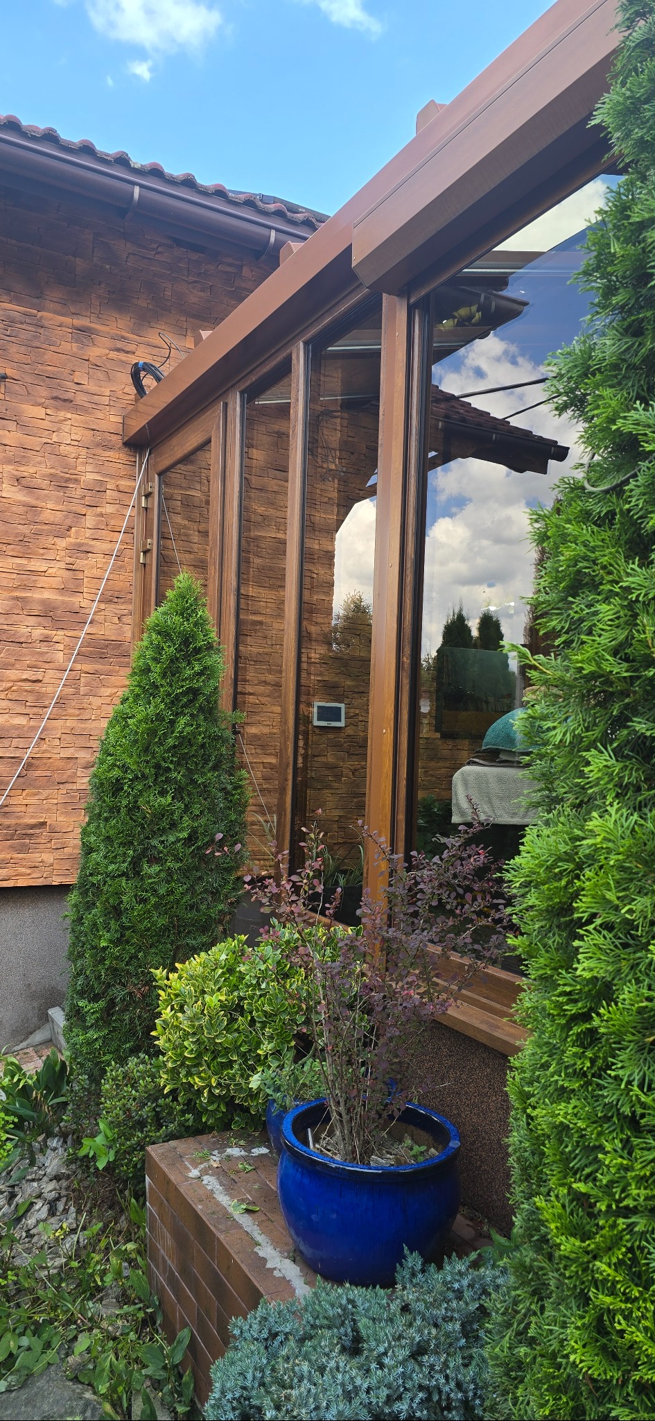 Zewnętrzny widok przeszklonej werandy z drewnianą konstrukcją, obok której rosną ozdobne krzewy i tuje w niebieskiej donicy. Na ścianie widoczny kamień elewacyjny.