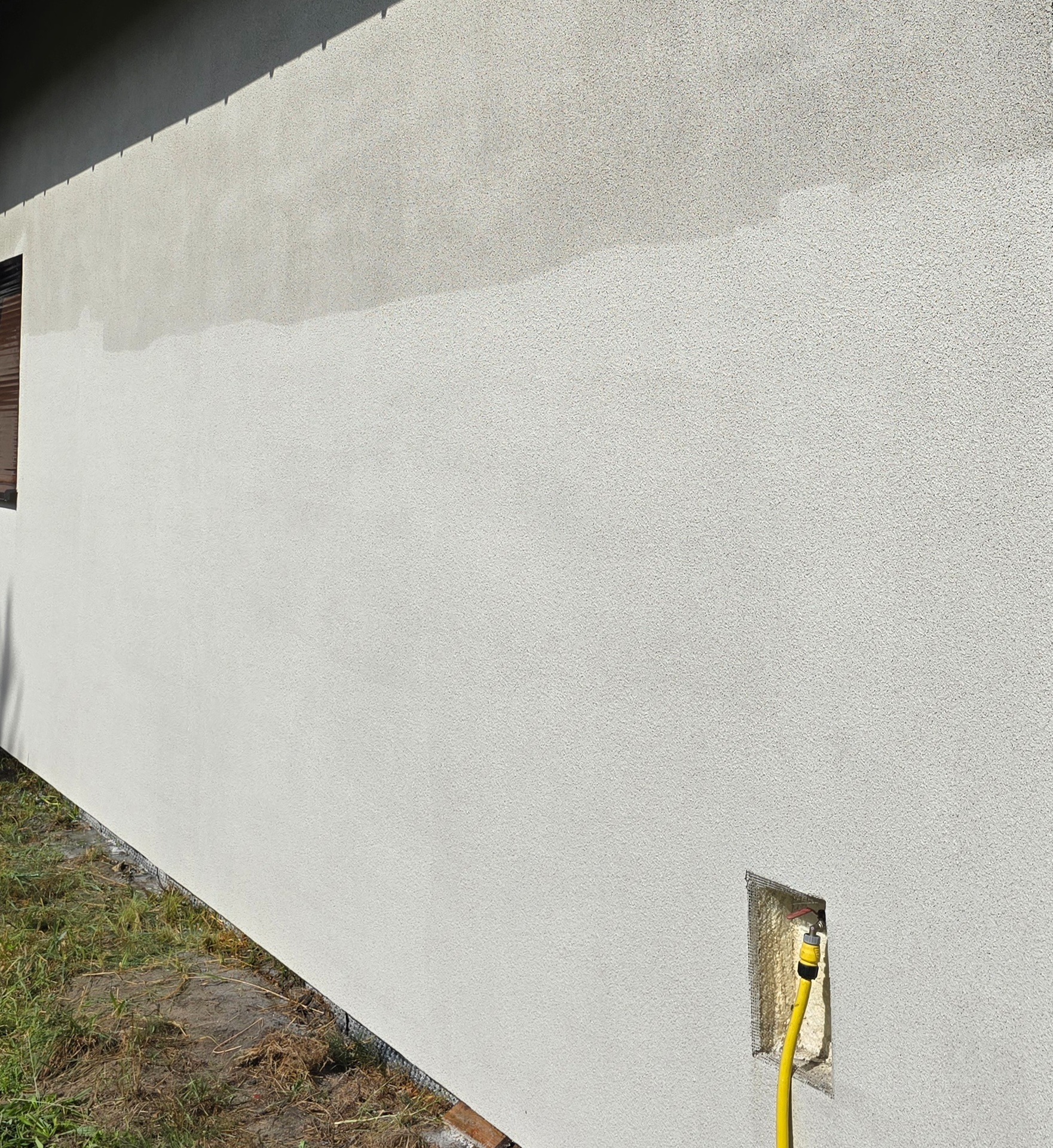 Ściana budynku z nową, jasną elewacją. Widoczny fragment okna, trawa przy budynku i żółty wąż w otworze w ścianie.