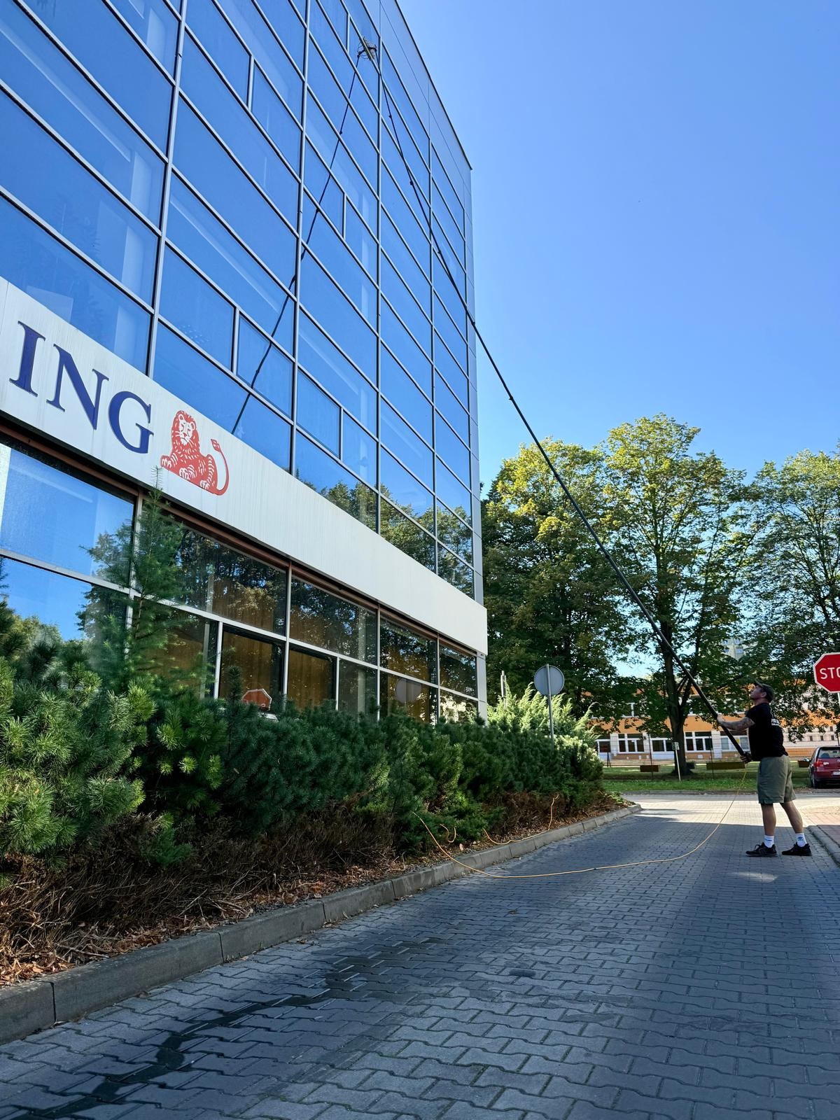 Mężczyzna myje okna biurowca ING za pomocą długiej lancy. Widoczne odbicia w szklanej fasadzie i znak stop na chodniku. Błękitne niebo w tle.