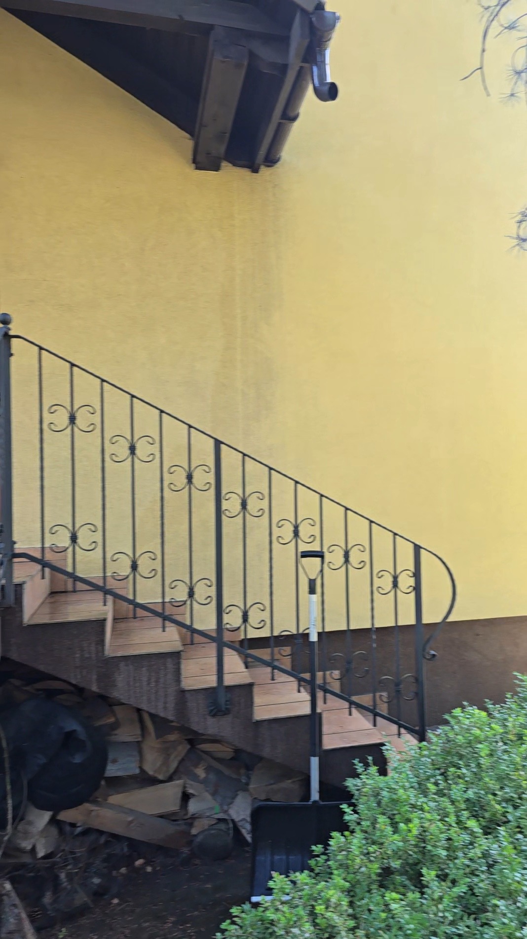 Żółta elewacja budynku z rynną, schody z kutą balustradą. Pod schodami drewno i łopata do śniegu. Fragment krzewu w prawym dolnym rogu.